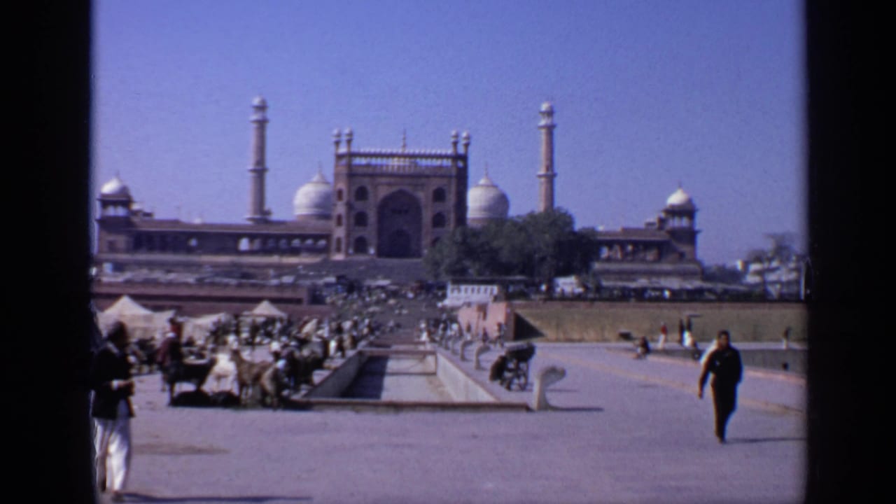 Wide courtyard view facing a domed mosque with minarets, people, carts, animals, and a long water channel.