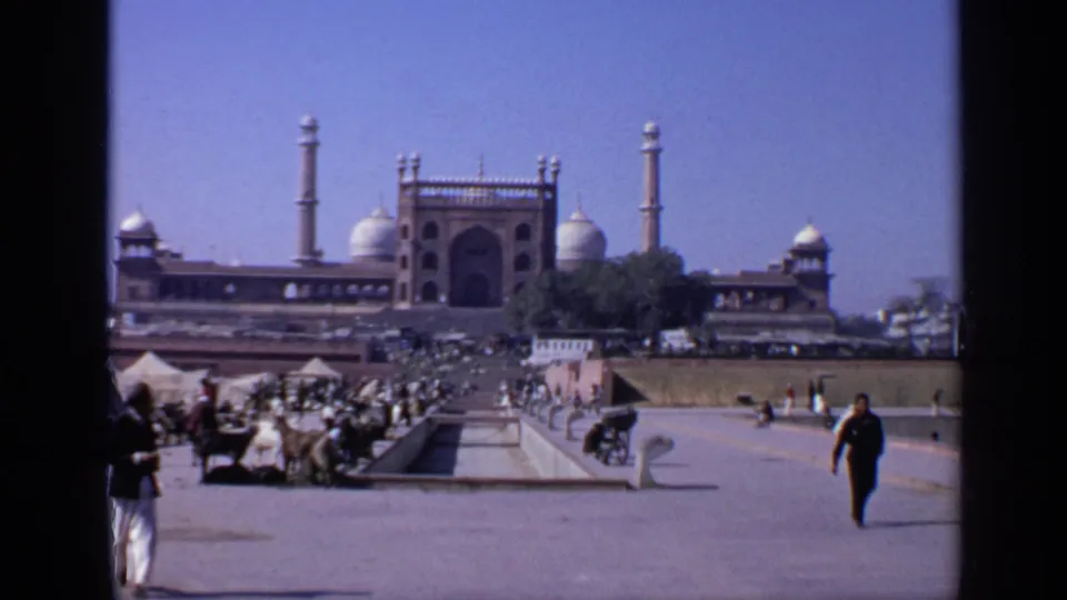 1970 8mm India home-movie frame showing a busy courtyard before a grand mosque with domes, minarets, visitors, carts, and animals.