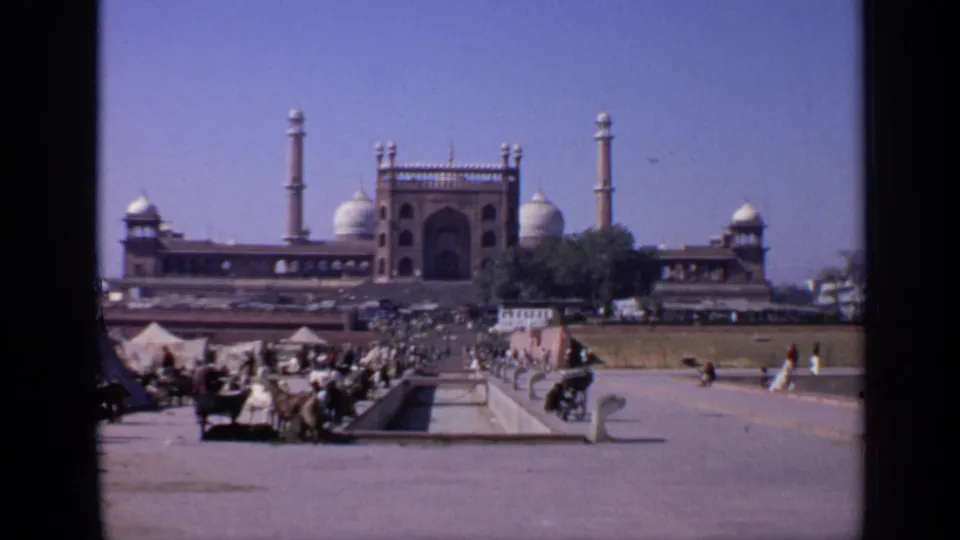1970 8mm India frame showing a public mosque courtyard with a water channel, visitors, animals, carts, domes, and minarets.