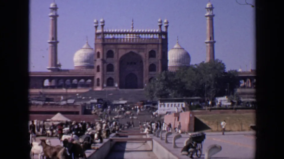 Closer 1970 India 8mm view of a mosque courtyard, with the arched facade, domes, minarets, visitors, animals, carts, and trees visible.
