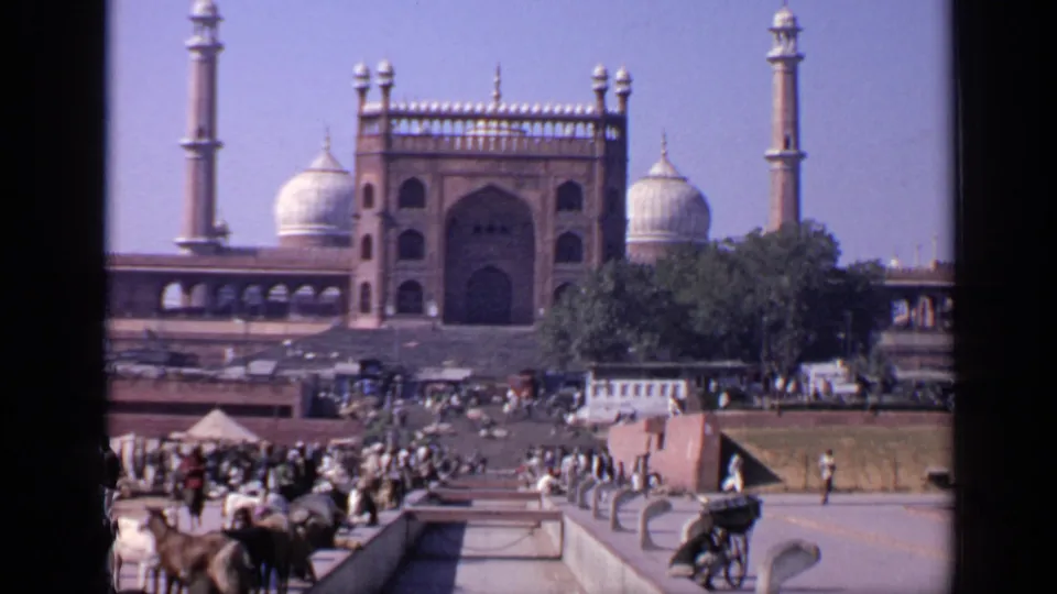 Wide 8mm frame of an Indian mosque complex in 1970, with domes, minarets, a tree-lined edge, and people crossing the plaza.