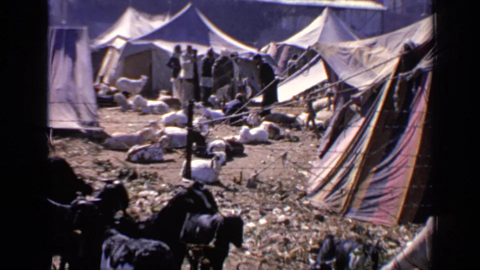 1970 8mm frame from India showing canvas tents, standing people, bundled materials, and shaded foreground figures near the mosque area.