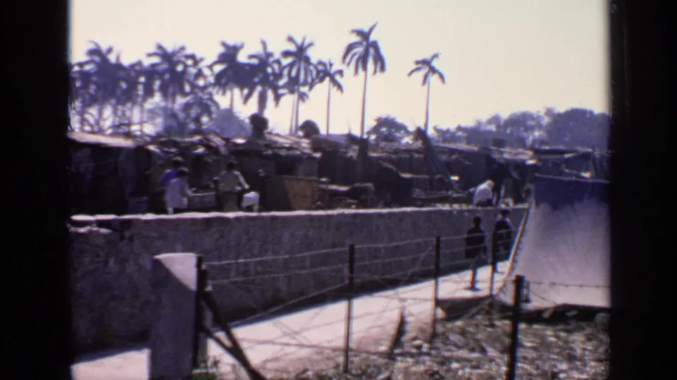 1970 8mm home-movie view in India of people walking along a paved path beside a wall, with palms and modest buildings beyond.
