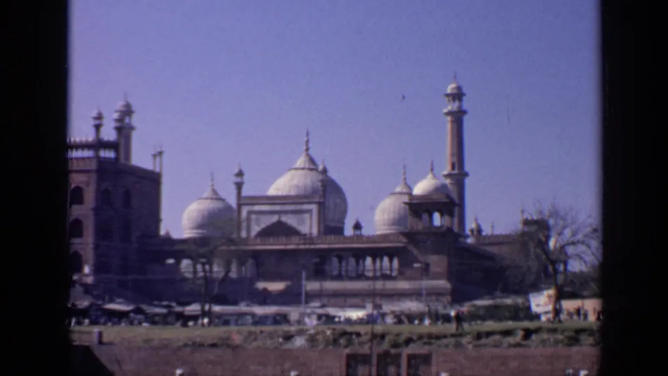 Wider 1970 India home-movie frame of the mosque complex with domes, minarets, open foreground, roadside activity, and clear sky.