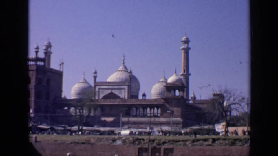 1970 India 8mm frame centered on the mosque's domes and tall minaret, with open ground, distant pedestrians, and a roadside sign.