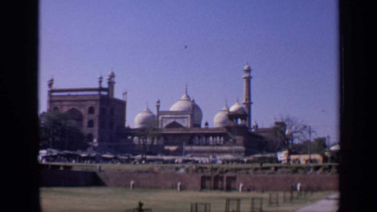 Wide view of the Taj Mahal complex across grass, wall, trees, stalls, and distant visitors.