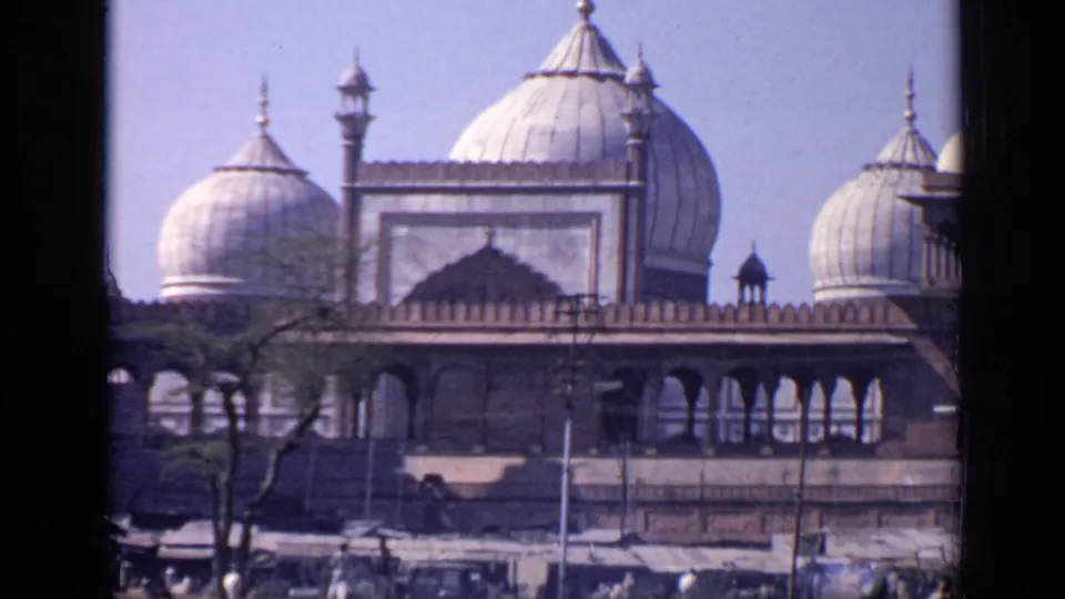 1970 India 8mm frame showing a domed mosque facade with arched colonnades, small towers, trees, and a dark window frame in the foreground.
