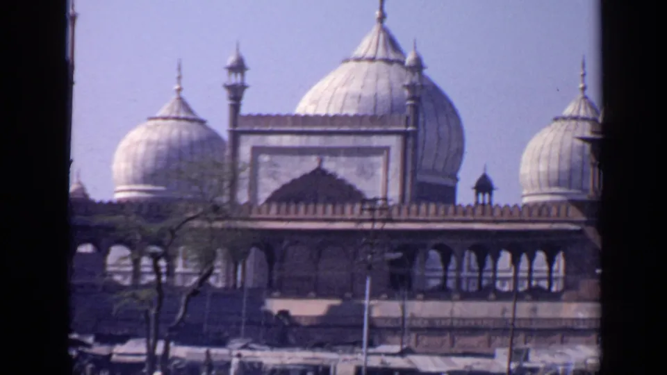 1970 India 8mm view of a mosque with large white domes, arched galleries, trees, and a dark vertical frame cutting through the scene.