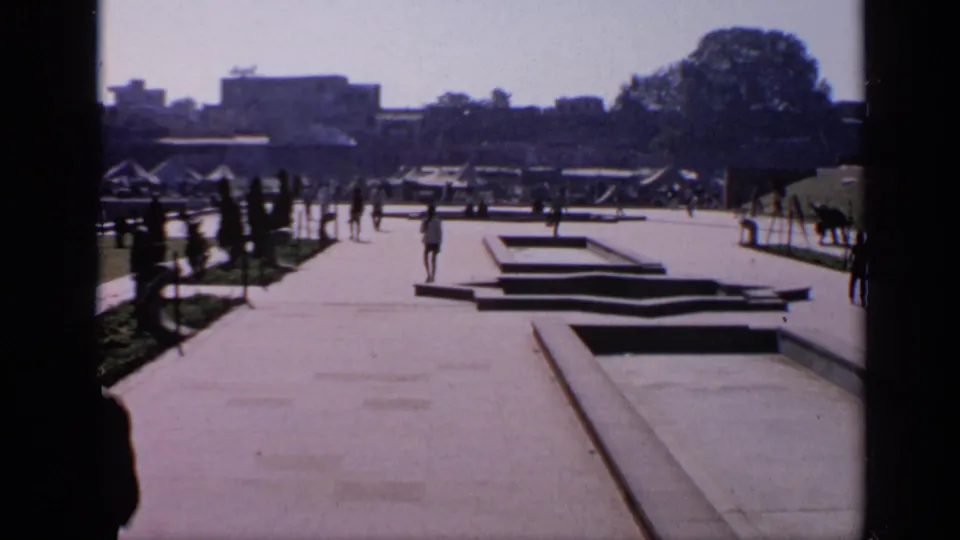 1970 India 8mm frame looking across a mosque courtyard with paved walkways, rectangular water features, trees, and distant pedestrians.