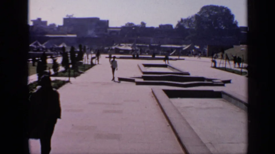 1970 India 8mm view across a mosque courtyard, with pedestrians on a paved path, rectangular basins, trees, and shaded foreground framing.