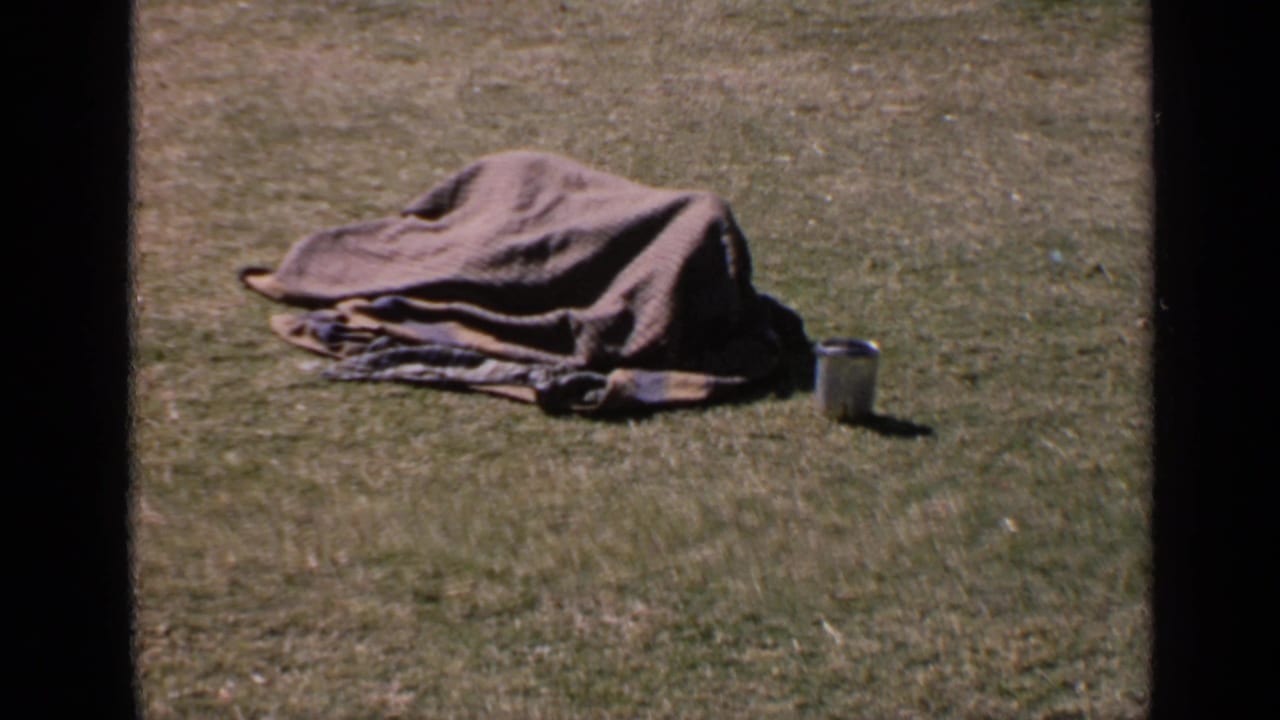 Person covered by a gray blanket lying on grass beside a small metal cup.
