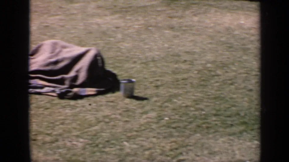 1970 India home-movie frame of a person covered by a gray blanket on a grassy field, with a metal cup beside them.