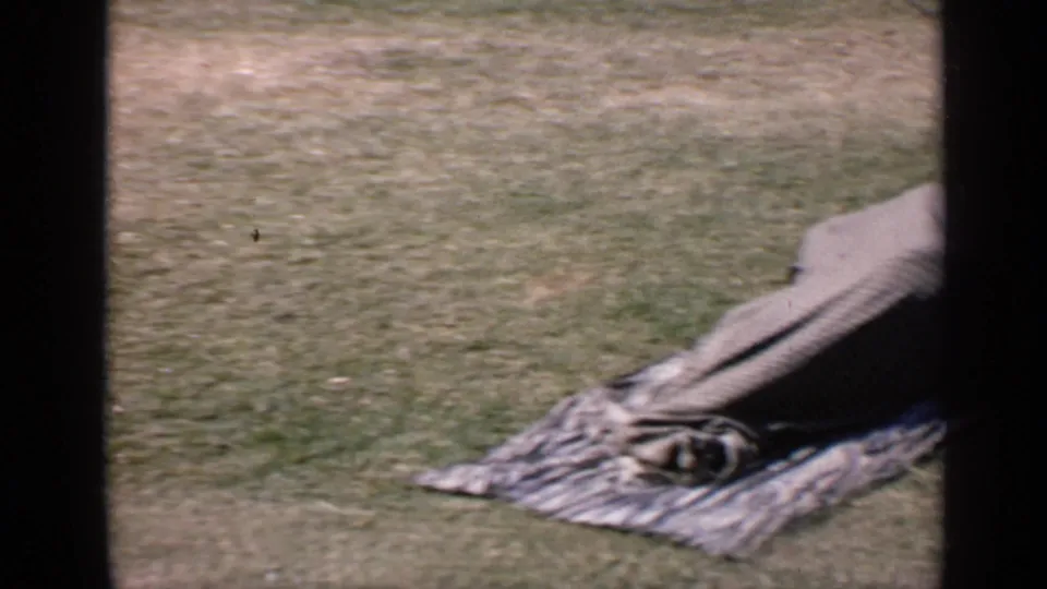 1970 India 8mm frame showing a blanket-covered person lying on a patterned cloth spread across grass.