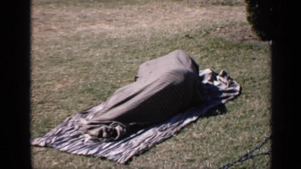 1970 India home-movie frame with a person resting under a gray blanket on a patterned cloth in bright outdoor light.