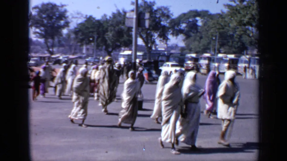 1970 India street view with pedestrians in loose garments and head coverings crossing a paved area near trees and parked vehicles.