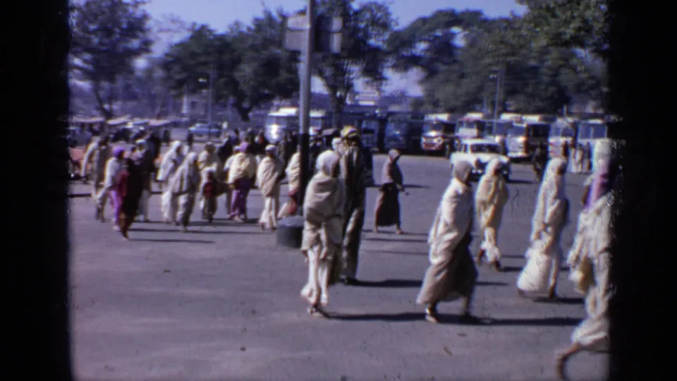 Handheld 8mm view of a public street in India, with people in traditional clothing walking past parked buses, cars, and trees.