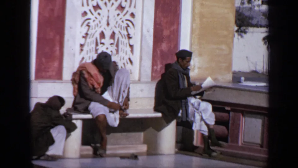 1970 India frame showing men seated and resting near a building with carved white wall detail and red and yellow surfaces.