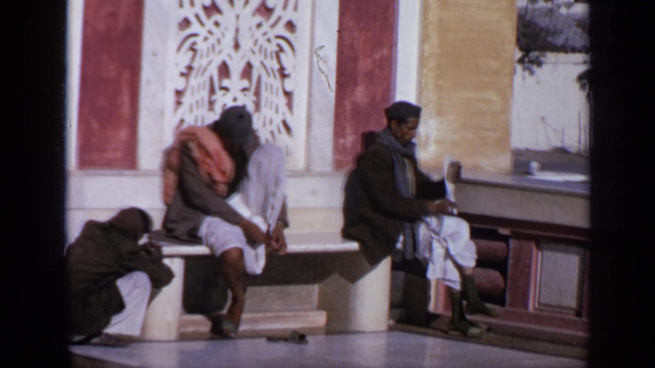 1970 India 8mm frame showing three men seated outside a temple-like building with carved red-and-white wall details.