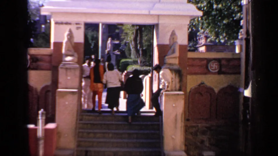 Visitors approach the top of a broad stairway in India, with carved walls and a visible symbol on the gate area.