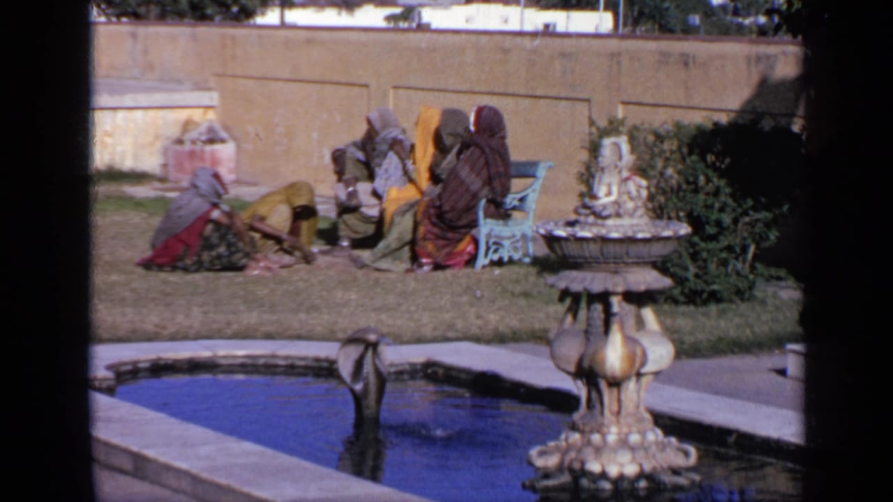 Women in sarees sit on a lawn near a fountain and pool in a sunlit courtyard.
