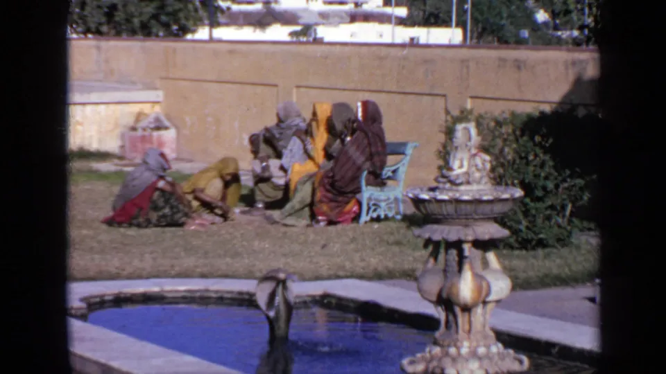 Women in sarees and head coverings sit together in a sunlit Indian courtyard near a fountain, pool, blue bench, and plaster wall.