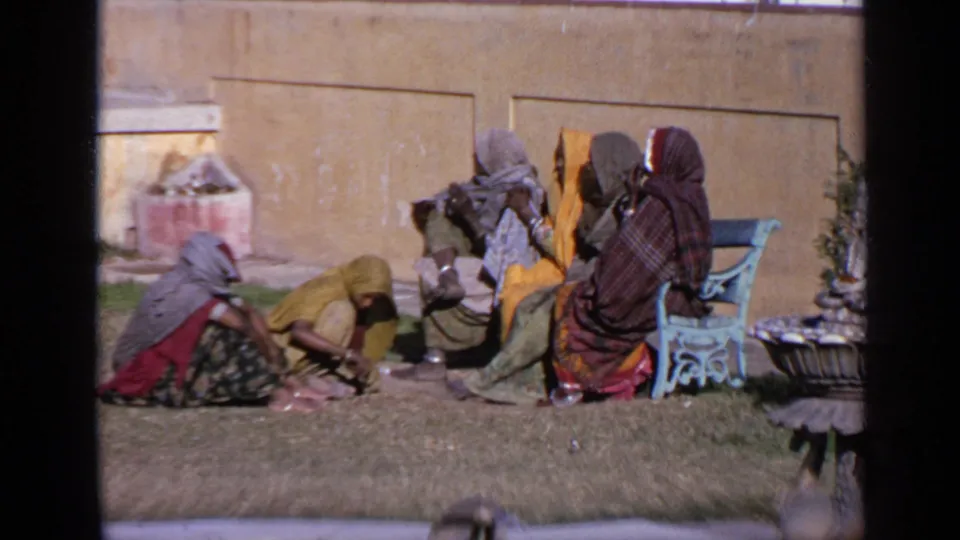 Closer 8mm frame of women in colorful sarees seated on grass in India, with a fountain base, wall, and blue bench nearby.