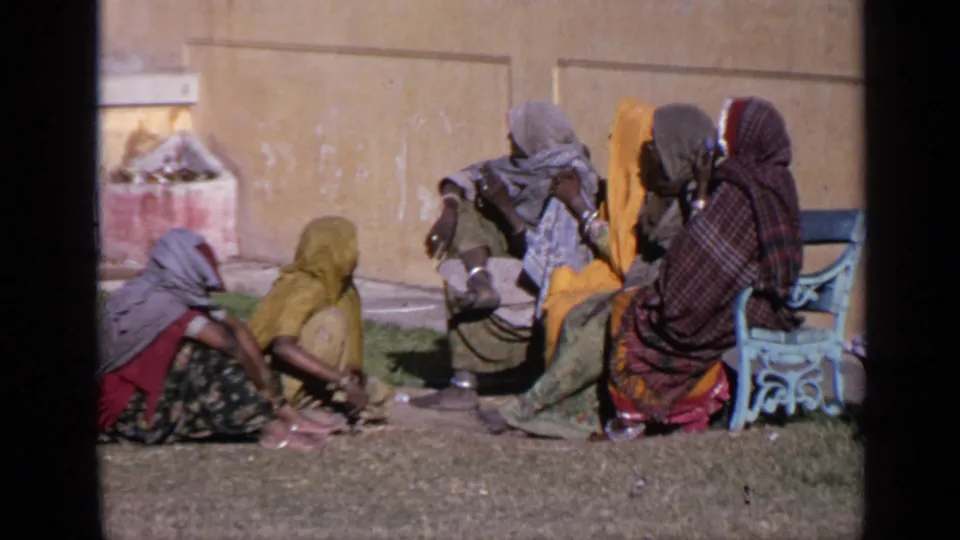 Close home-movie view of Indian women in sarees and head coverings sitting together on a lawn beside a blue bench and courtyard wall.
