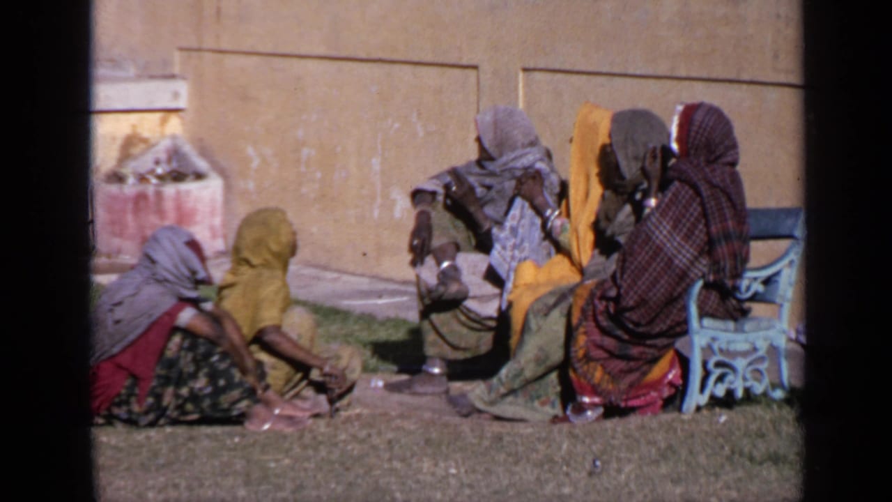 Women in headscarves sit on grass and a blue bench beside a yellow wall.