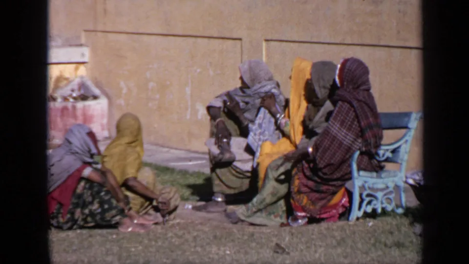 Women appear seated in conversation outside a temple in India, with headscarves, grass, a yellow wall, and a blue bench visible.