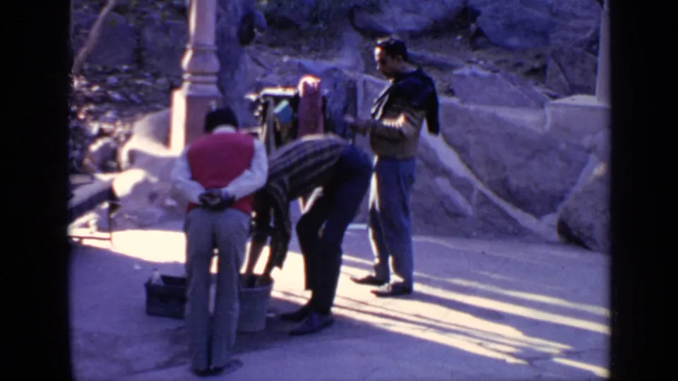 1970 India 8mm frame of men near a stone temple-area structure, one bending toward a bucket or basin while others stand nearby.