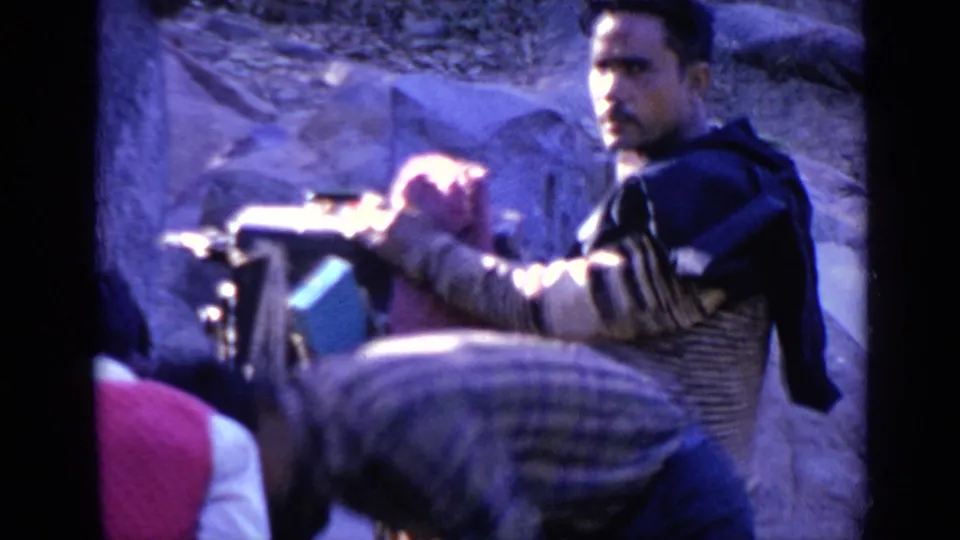 Closer 1970 India 8mm view of a man near a bucket or basin in a temple-area setting, with another figure bent forward in the foreground.