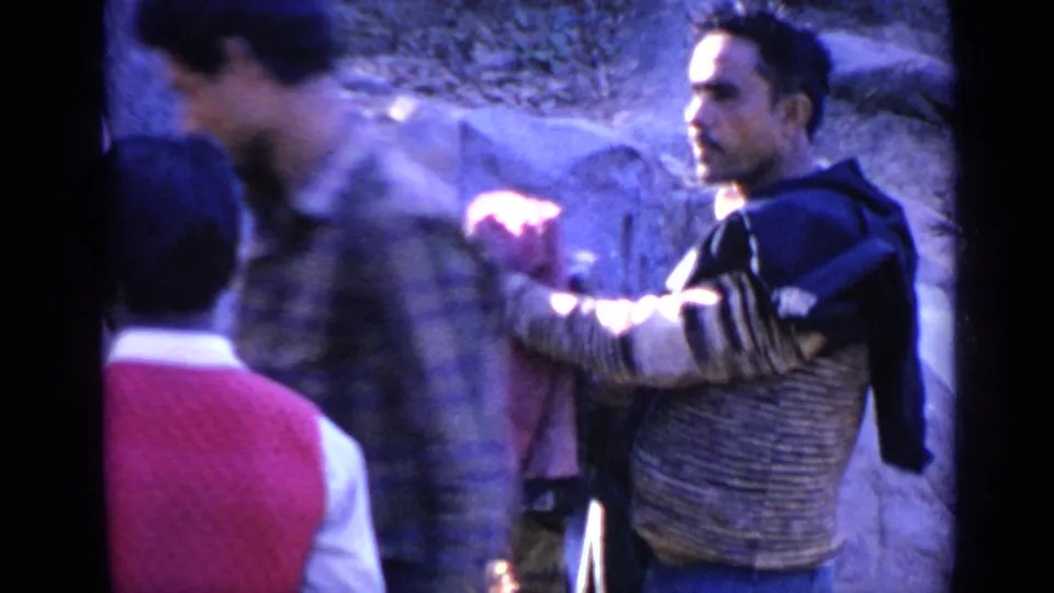 1970s India home-movie frame with a blurred man in plaid crossing the foreground and another adult looking toward the camera area.