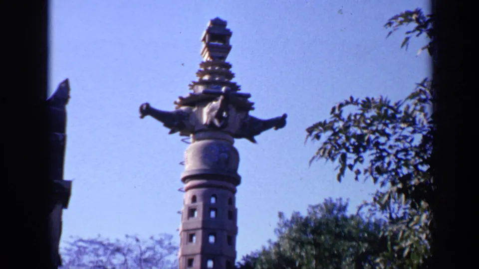 1970s India 8mm view of a tall ornate stone tower filmed from below, framed by blue sky, trees, and dark film edges.