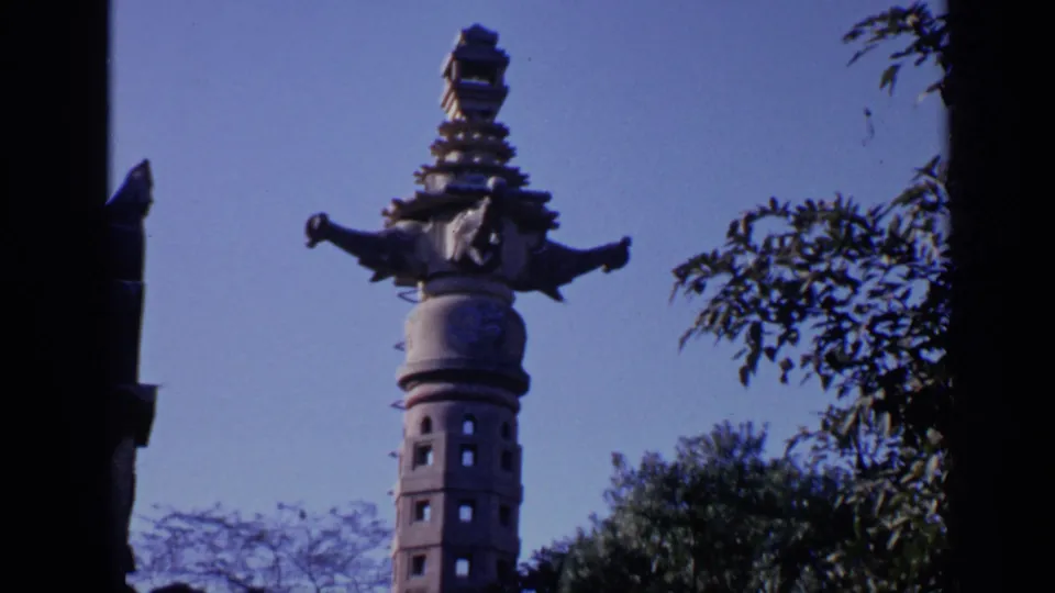 1970s India home-movie frame showing the stone tower from a low angle, with tree branches and a clear blue sky around it.