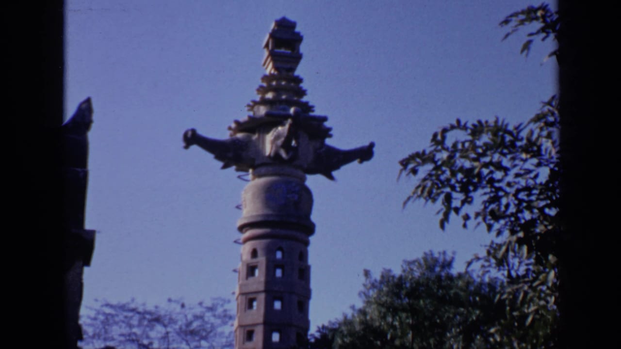 Temple grounds with a decorative column, carved tower, trees, and blue sky.