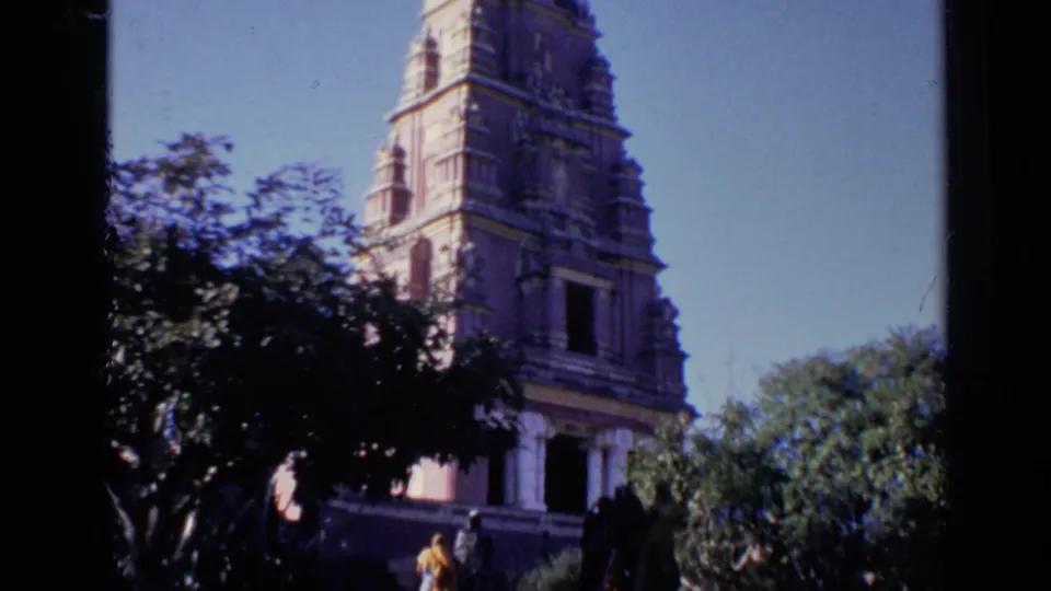 1970 India 8mm frame of a carved temple tower behind trees, with visitors visible near the lower entrance area.