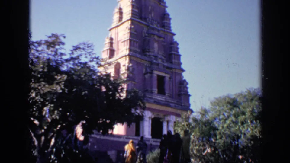 1970 India 8mm frame showing a temple entrance area, carved tower, trees, and several visitors in partial shade.