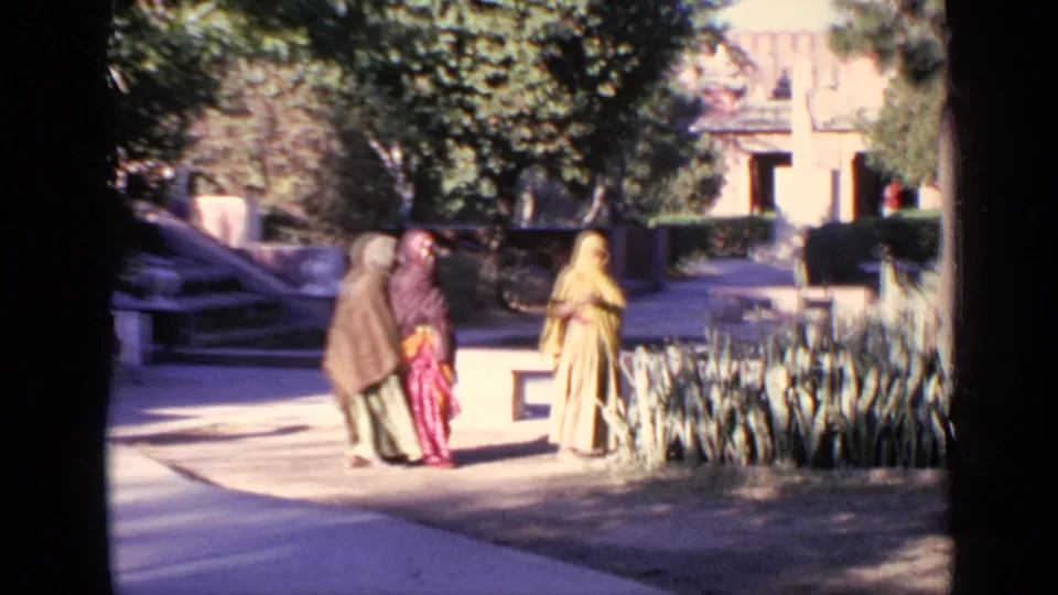 1970 India 8mm frame of three women in saris or head coverings walking beside plants and steps in a temple courtyard.