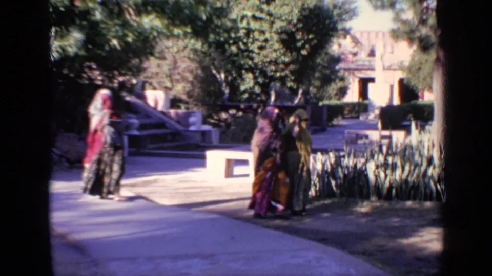 1970 India 8mm frame of women walking across a temple courtyard path with trees, plants, and pale structures behind them.