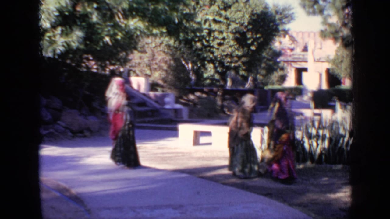 Women in traditional clothing walk along a sunlit path near trees and low walls.