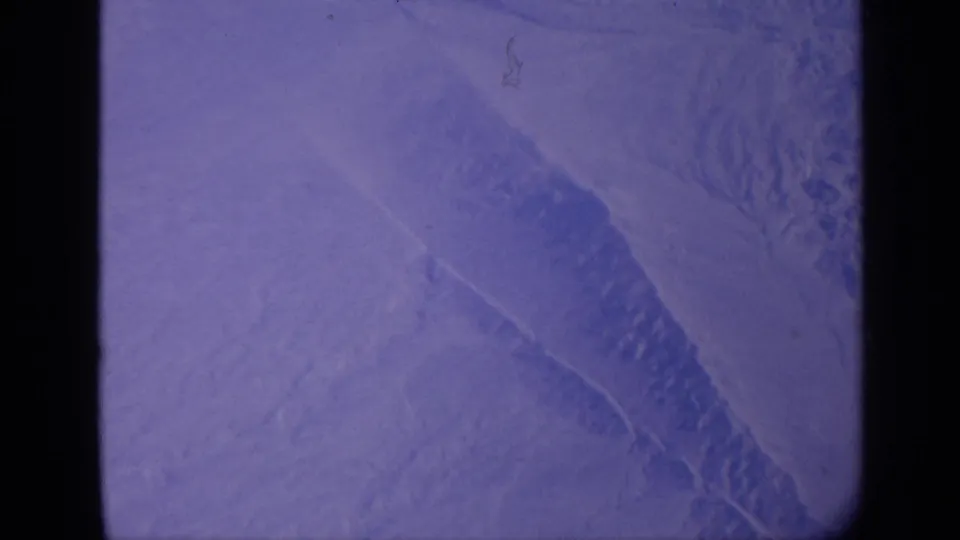 1970 India home-movie frame of a snow-covered slope with a darker diagonal ridge, faded blue-purple color, and film dust.