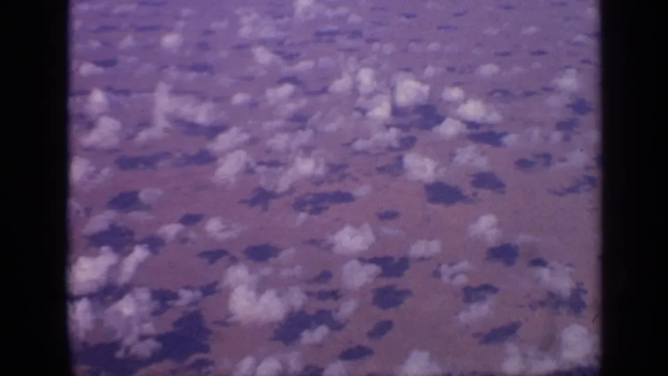 1970 home-movie aerial footage over India with small clouds scattered across a hazy surface, framed by dark 8mm film edges.
