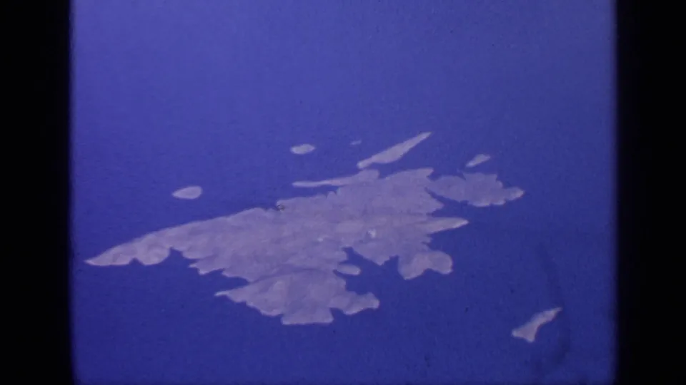 Aerial passenger-window view in 1970 8mm footage showing pale islands surrounded by blue water, with film grain and color shift.