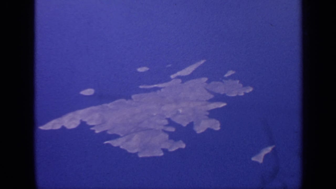 1970 India 8mm aerial view of a pale treeless island shape surrounded by blue water and film gate edges.