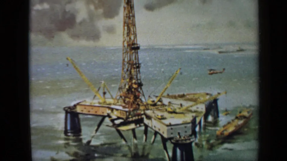 1970 United Kingdom 8mm frame of a painted offshore rig scene, with a small vessel alongside and cloudy sky in the background.