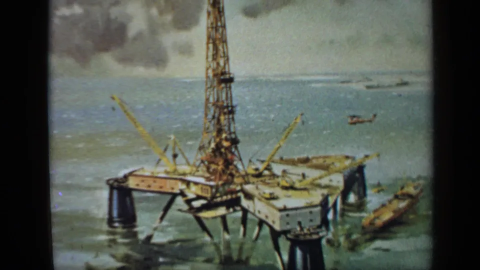 1970 United Kingdom 8mm frame showing an offshore drilling platform with cranes, a tall derrick, choppy water, and a helicopter nearby.