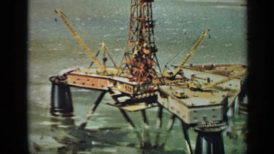Closer 1970 United Kingdom 8mm view of the offshore platform deck, derrick base, cranes, and surrounding green-blue sea.