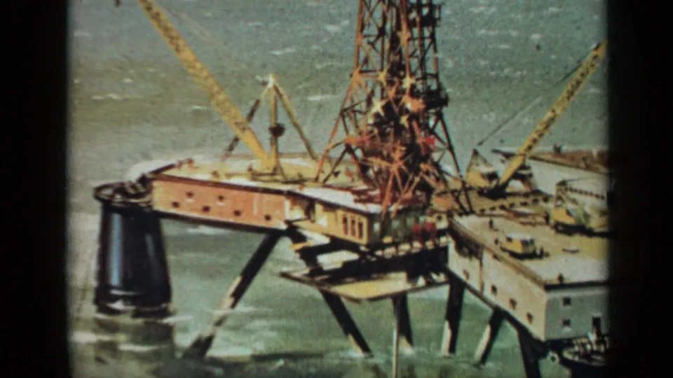 Final close 8mm frame of the 1970 United Kingdom offshore drilling platform, centered on the derrick, cranes, deck, and support legs over the sea.