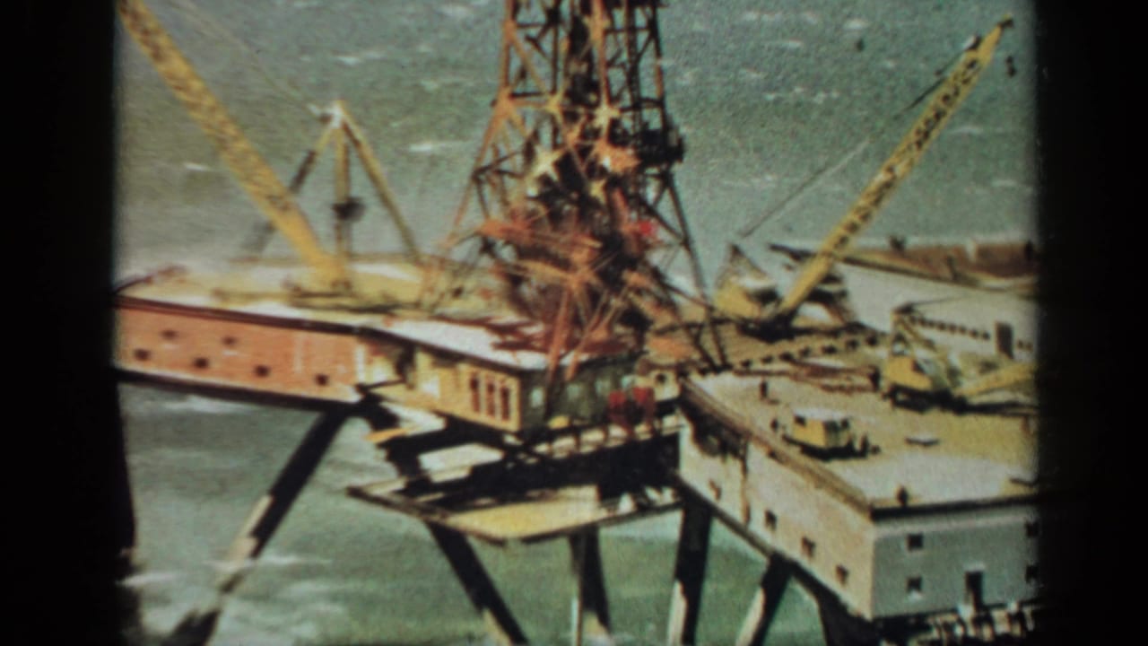 Offshore platform deck with cranes, central tower, workers, and surrounding water.