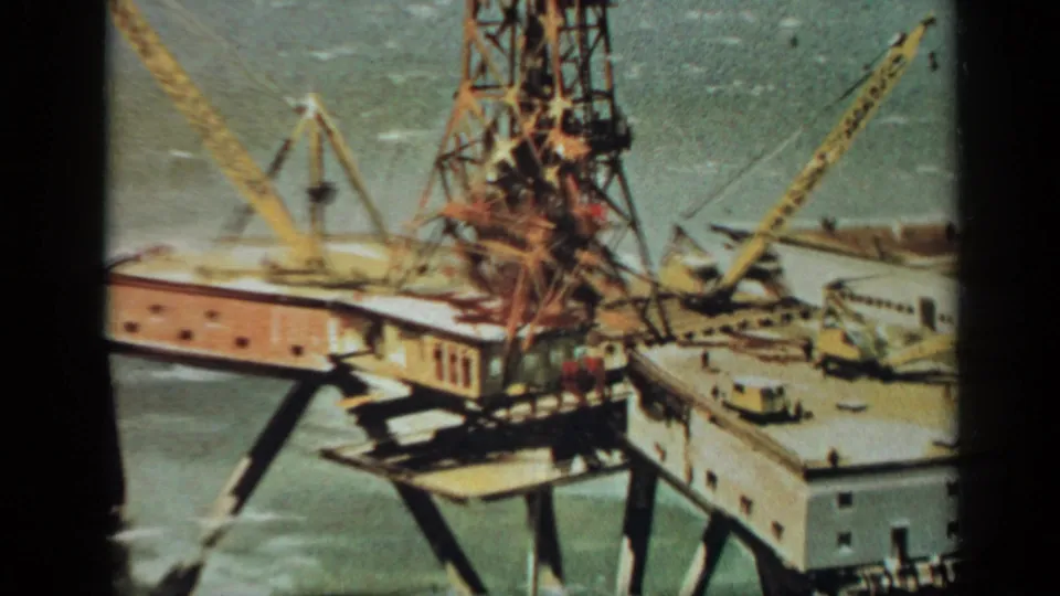 1970 United Kingdom 8mm frame showing an offshore industrial platform with yellow cranes, a derrick-like tower, workers, and sea around it.